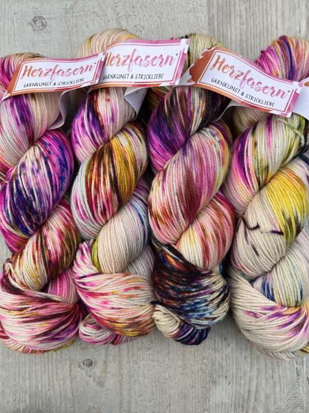 Vier handgefärbte Stränge Wolle in bunten Farben – Pink, Gelb, Orange, Blau und Sprenkel – liegen nebeneinander auf hellem Holz. Das Herzfaser-Logo ist auf den Etiketten sichtbar.